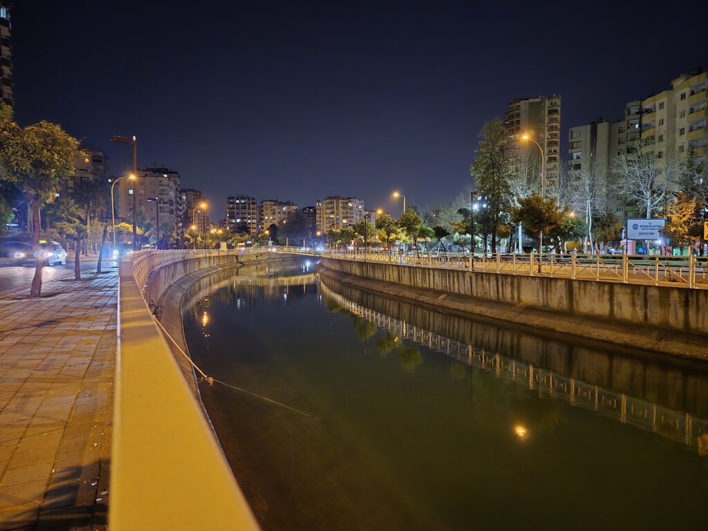 Gece çekimleri ile aydınlatılmış bir şehir manzarası; su kenarında yürüyüş yolu, binalar ve sokak lambaları.