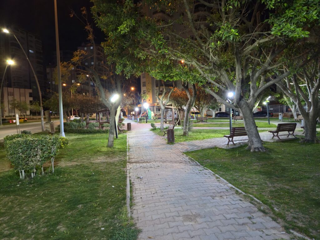 Gece çekimi yapılan bir parkın görünümü, yeşil ağaçlar ve banklarla dolu bir yürüyüş yolu, sokak lambalarının aydınlattığı alan.