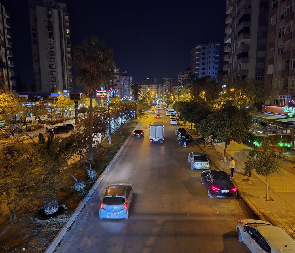 Geceleri aydınlatan sokak ışıkları ve binalar arasında hareket eden araçlar, gece manzarası.