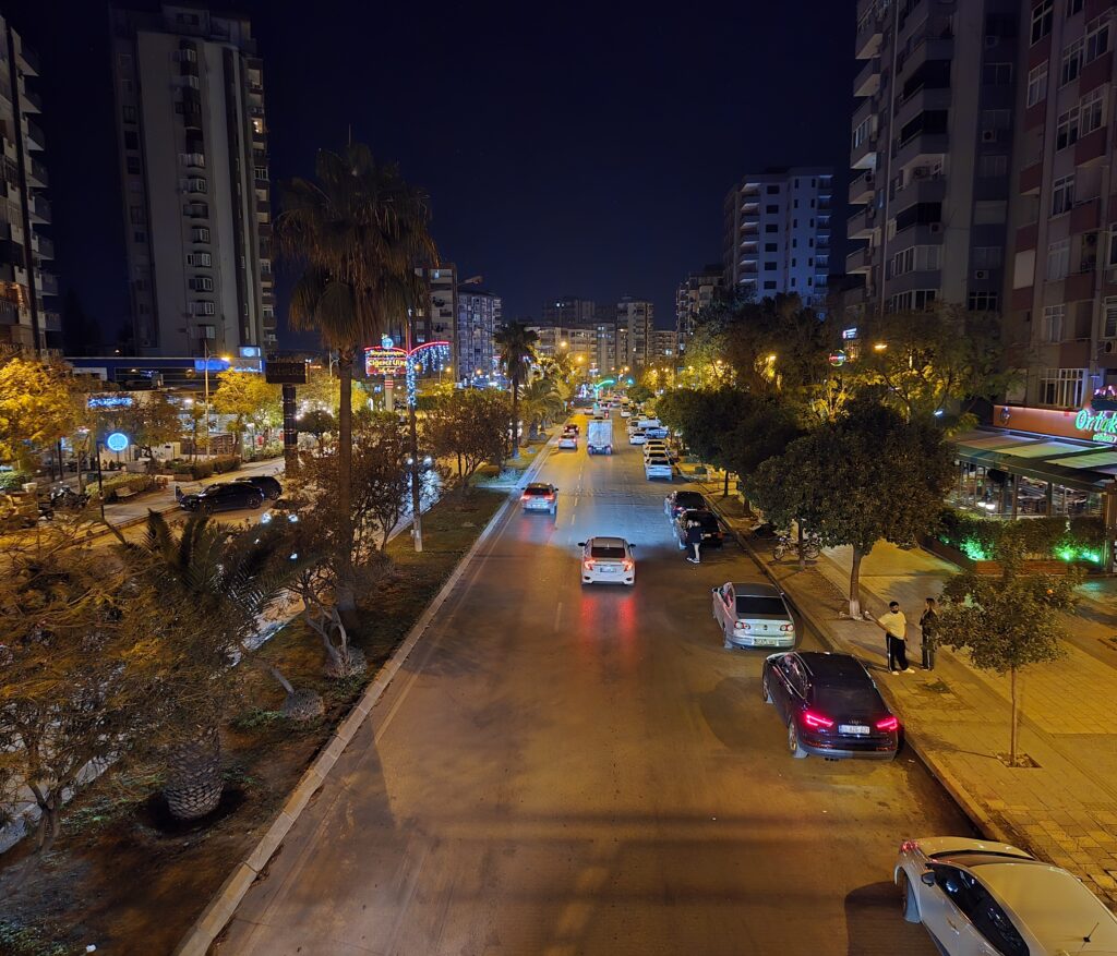 Gece çekimi, aydınlatılmış bir cadde, araçlar ve palmiyelerle çevrili bir şehir manzarası.